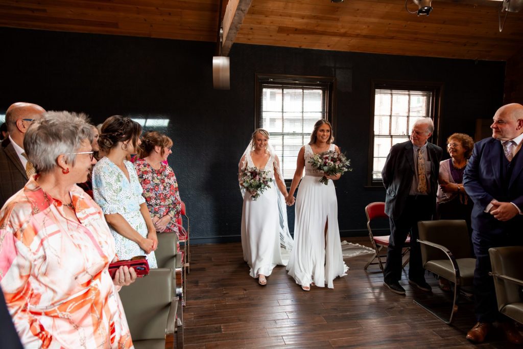 The bride & bride walking hand in hand up the aisle together at The Chimney House wedding venue, Kelham Island, Sheffield. Isabelle B Photography
