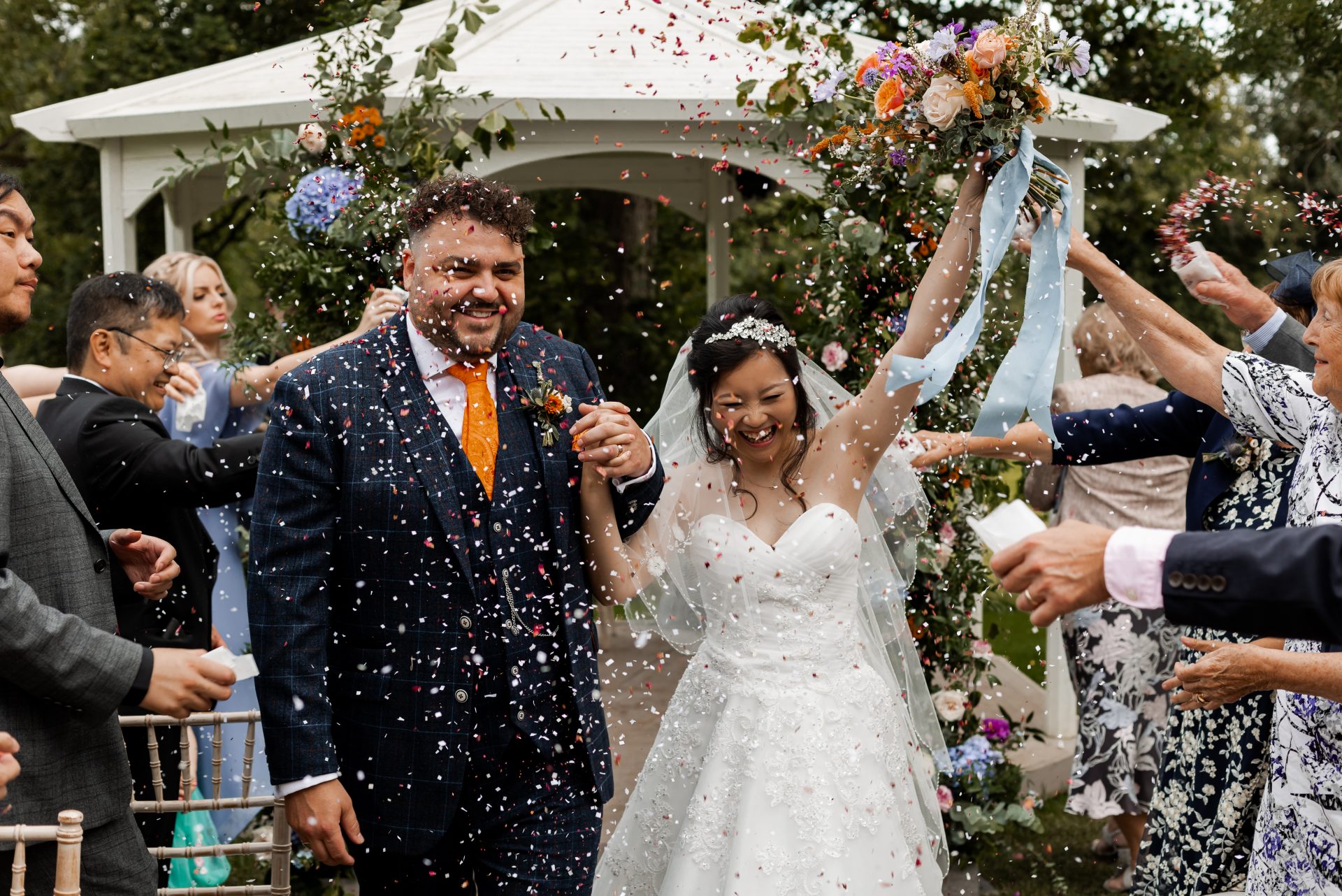 Newly married Bride & Groom walk down the aisle as confetti is thrown. The bride is ecstatic and raises her bouquet in the air