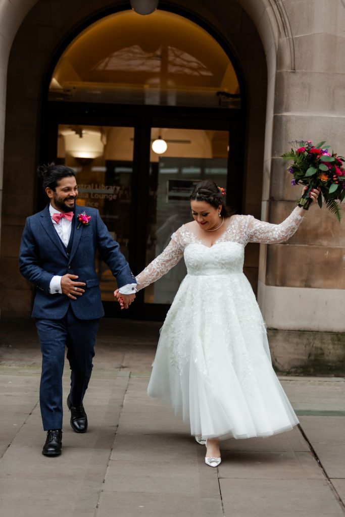 Husband and wife strolling together and laughing. The bride is wearing a white midi-wedding dress and waving her bouquet in the air