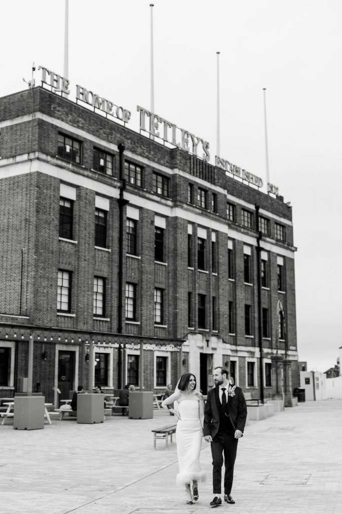 Black and white photograph of a newly married couple walking hand in hand with the icon Tetley, Leeds building in the background 