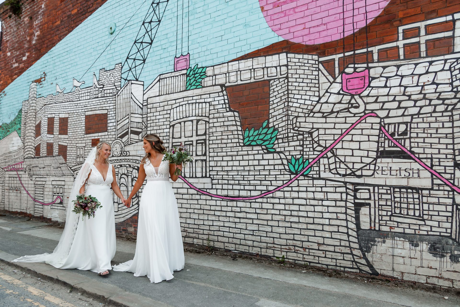 Bride & Bride walk together hand in hand in front of the Jo Peel mural at Kelham Island. Isabelle B Photography, Sheffield Wedding Photography