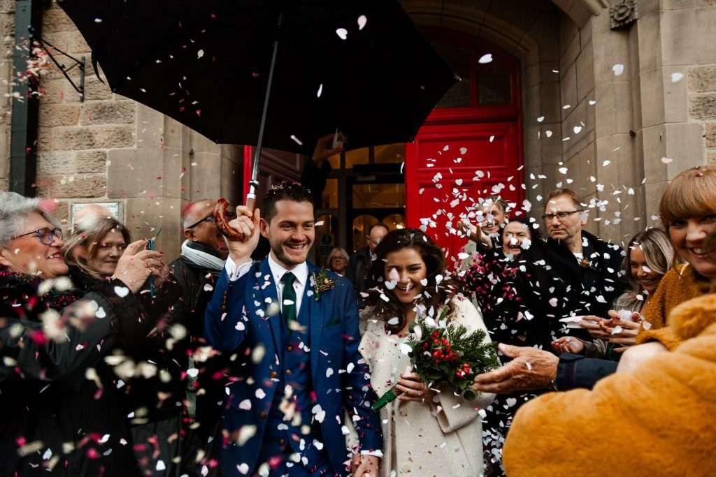 Groom & bride walking out of the Bakewell registry office holding a black umbrella and being covered with confetti