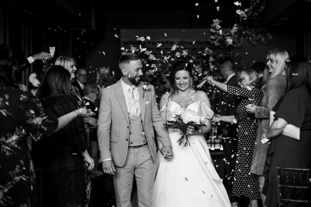 Bride and groom walking pack up the aisle and confetti is being thrown. Inside the ceremony room at the Manor House, Lindley. Black & White