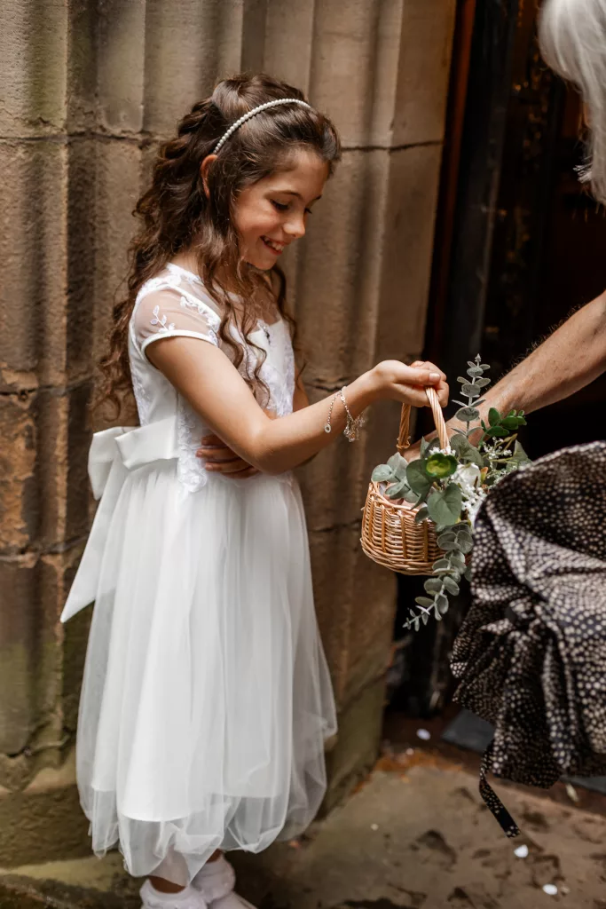A flower girl holding a basket of confetti outside the church. A lady leans in to grab a handful of confetti