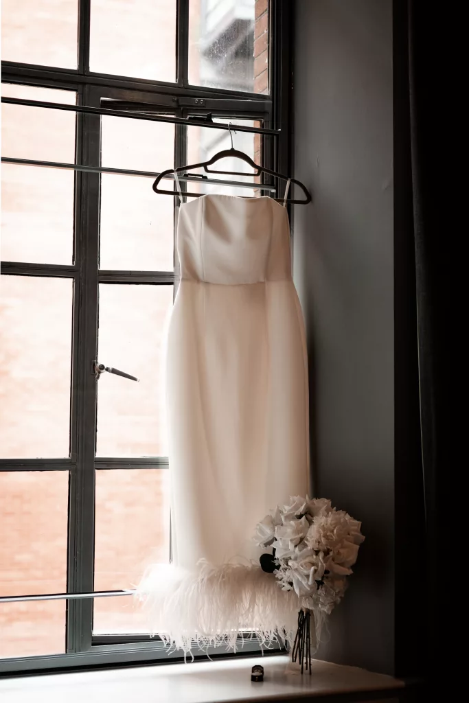 A shot of the wedding dress hung in the hotel window with the flowers and engagement ring