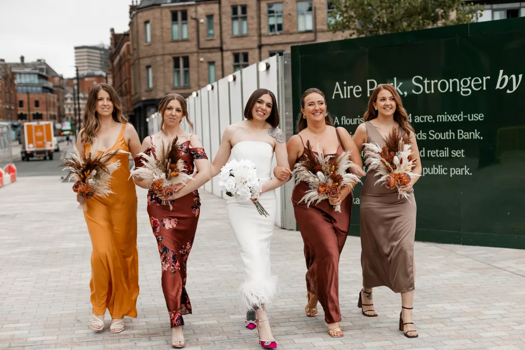 The bride and her bridesmaids walking together towards the wedding venue, The Tetley, Leeds. The bride wearing an alternative three quarter length wedding dress and the bridesmaids wearing a variety of dresses all of earthy tones - Isabelle B Photography - Wedding Photography