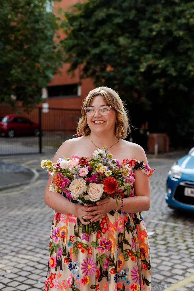 The bride outside her wedding venue before the wedding ceremony. The bride is wearing a colourful flower embroidered wedding dress and holding a colour flower bouquet