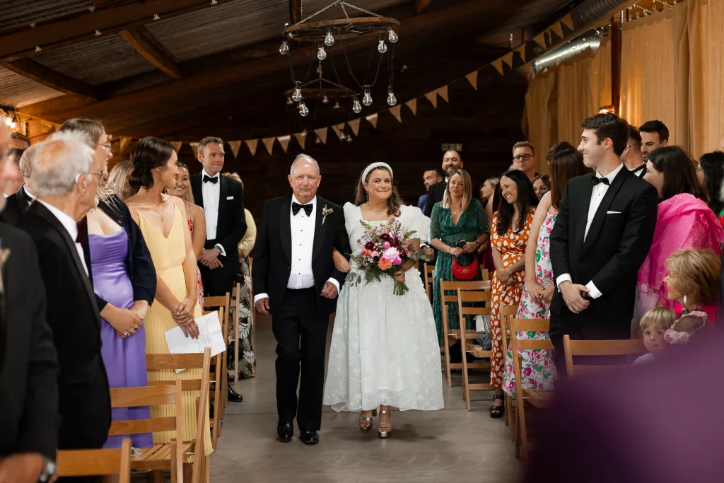 The bride walking down the aisle arm in arm with her father at The Grange Barn, Cheshire. The bride is wearing an alternative wedding dress