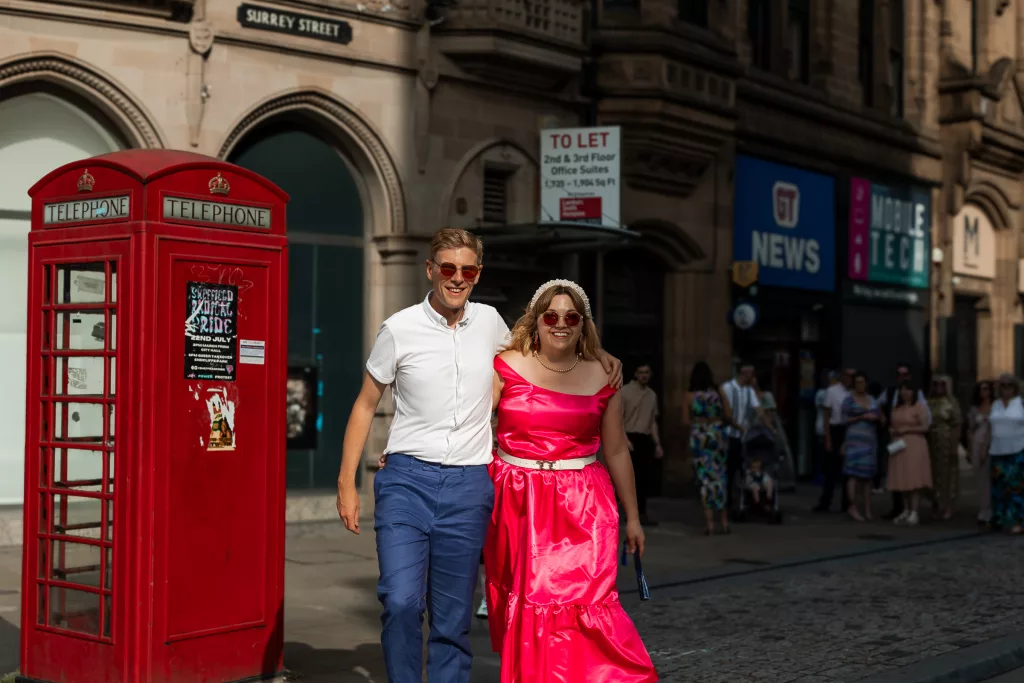 The soon to be bride & groom walking together outside Sheffield Town Hall. The bride is wearing an alternative bright pink wedding dress - Isabelle B Photography