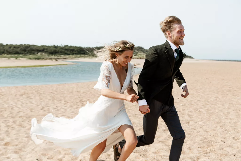 Newly married Bride & Groom running on the beach at Harvest Moon wedding venue in Scotland. The bride is wearing a rembo styles wedding dress and flower crown