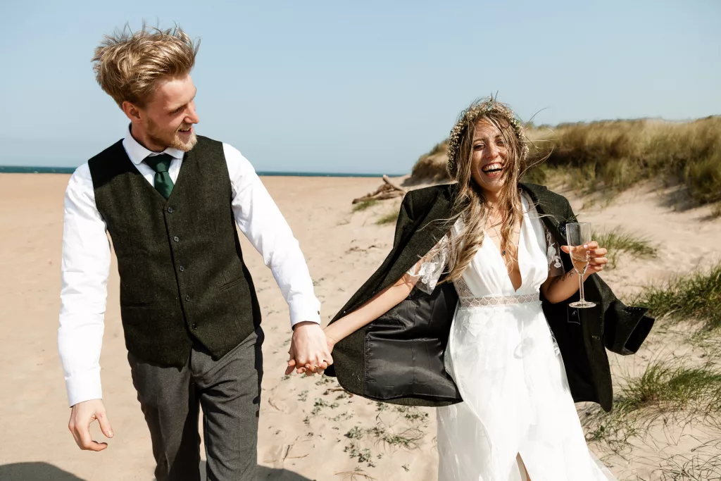 Bride & groom walking together happily on the beach in Dunbar, Scotland. The bride is wearing a lace Rembo Styles wedding dress, flower crown, and her new husbands suit jacket draped over her shoulders
