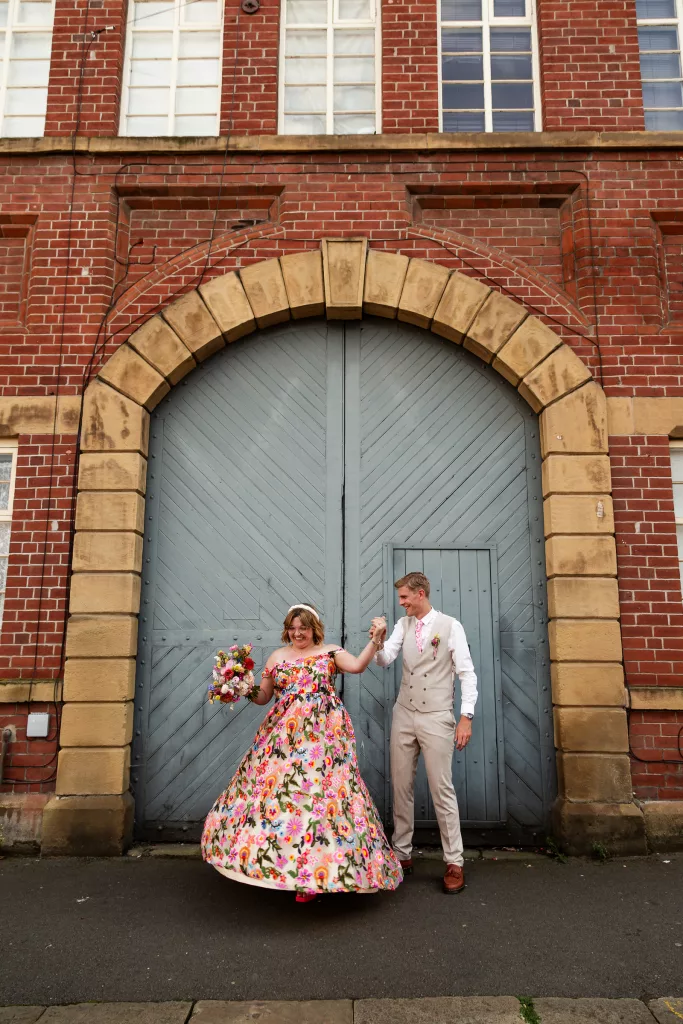 Newly married husband and wife pictured in front of a big blue door as the groom twirls his wife. The bride is wearing a colourful flower embroidered dress and groom is wearing a light coloured suit with a pink tie to match the brides dress
