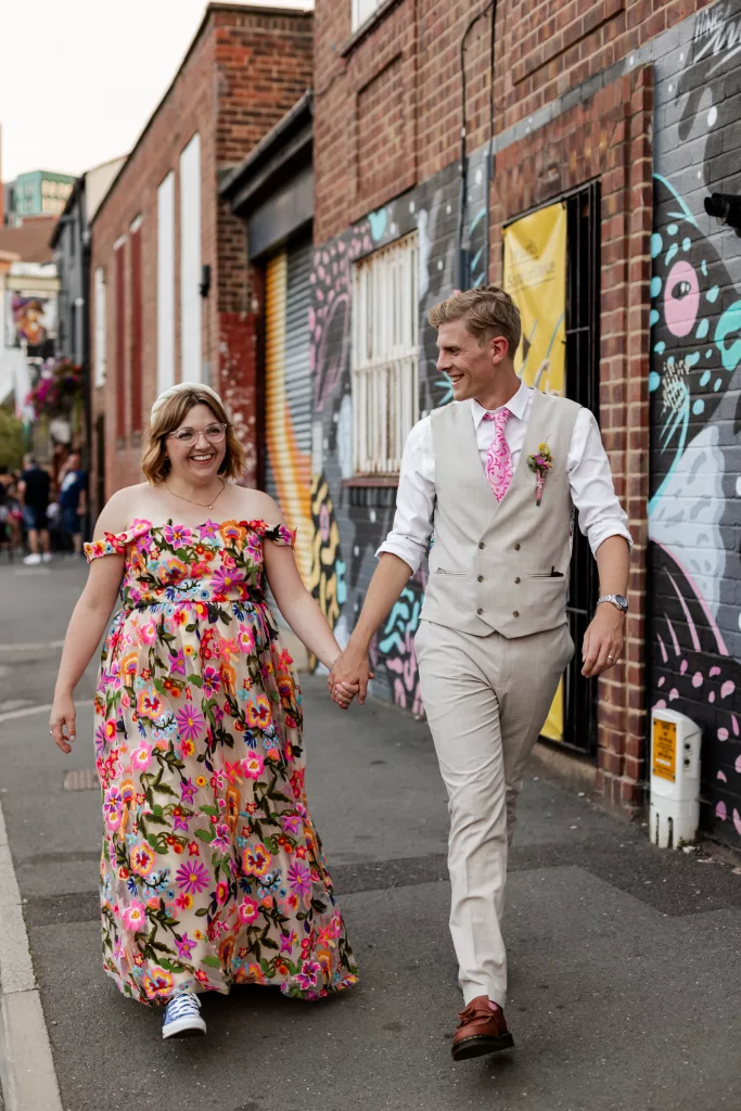 Newly married husband and wife walking together. The bride is wearing a colourful flower embroidered dress and groom is wearing a light coloured suit with a pink tie to match the brides dress