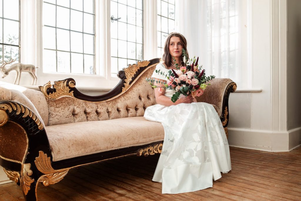 Wide shot of the bride sat on a ches long sofa holding her flower bouquet before her wedding ceremony at Whirlowbrook Hall - Isabelle B Photography