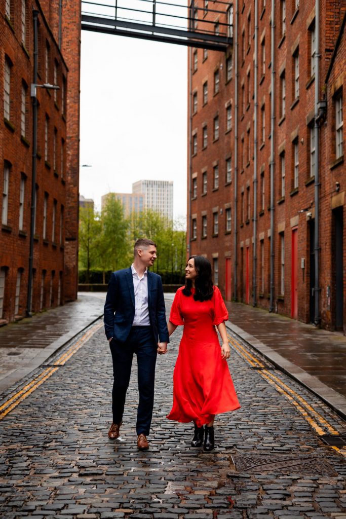 Newly engaged couple walking together in Ancoats, Manchester - The bride to-be is wearing a vivid red dress & the groom to-be is wearing a blue suit - Engagement Shoot - Isabelle B Photography