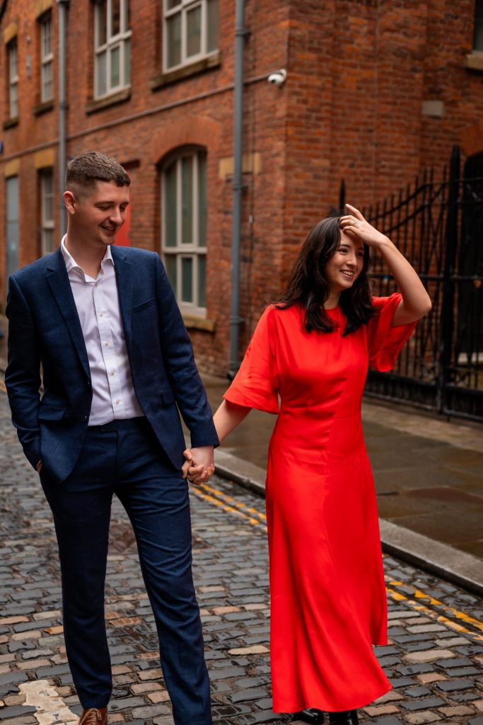 Newly engaged couple walking together in Ancoats, Manchester - The bride to-be is wearing a vivid red dress & the groom to-be is wearing a blue suit - Engagement Shoot - Isabelle B Photography