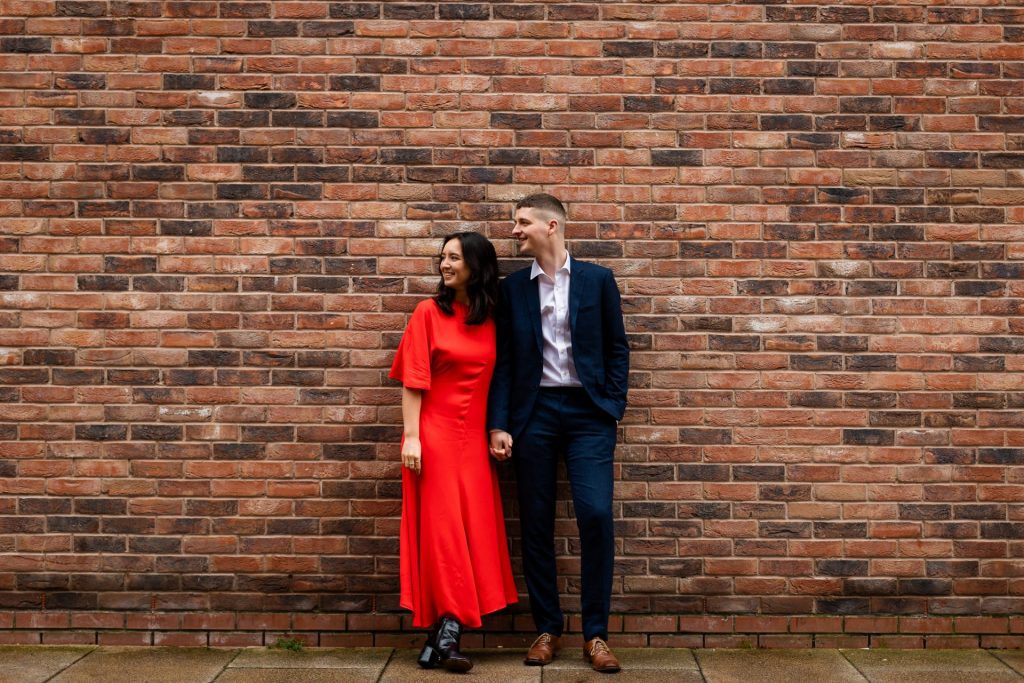 Engaged couple stood together leaning against a tall brick wall in Ancoats, Manchester - The bride to-be is wearing a vivid red dress, and the groom to-be is wearing a blue suit - Engagement Shoot - Isabelle B Photography