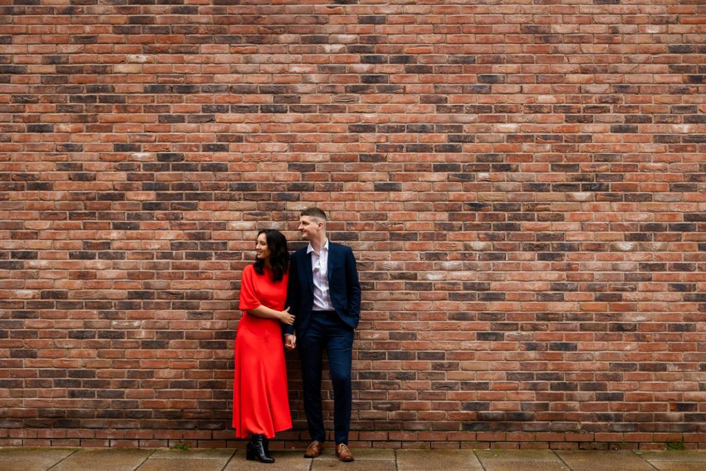 Engaged couple stood together leaning against a tall brick wall in Ancoats, Manchester - The bride to-be is wearing a vivid red dress, and the groom to-be is wearing a blue suit - Engagement Shoot - Isabelle B Photography