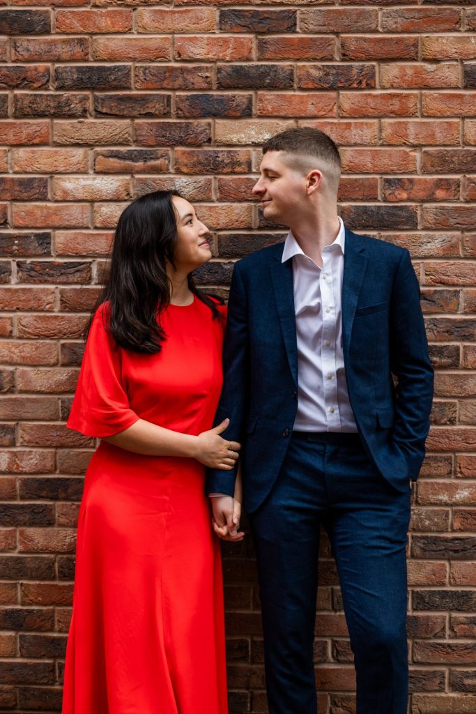 Engaged couple stood together leaning against a tall brick wall in Ancoats, Manchester - The bride to-be is wearing a vivid red dress, and the groom to-be is wearing a blue suit - Engagement Shoot - Isabelle B Photography