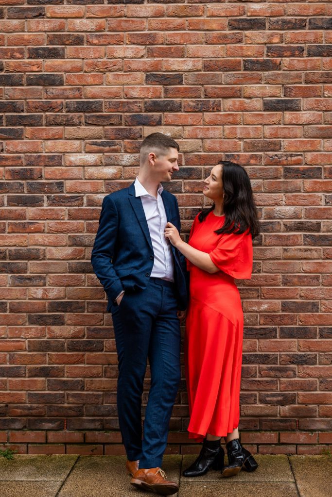 Engaged couple stood together leaning against a tall brick wall in Ancoats, Manchester - The bride to-be is wearing a vivid red dress, and the groom to-be is wearing a blue suit - Engagement Shoot - Isabelle B Photography