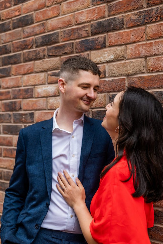 Engaged couple stood together leaning against a tall brick wall in Ancoats, Manchester - The bride to-be is wearing a vivid red dress, and the groom to-be is wearing a blue suit - Engagement Shoot - Isabelle B Photography