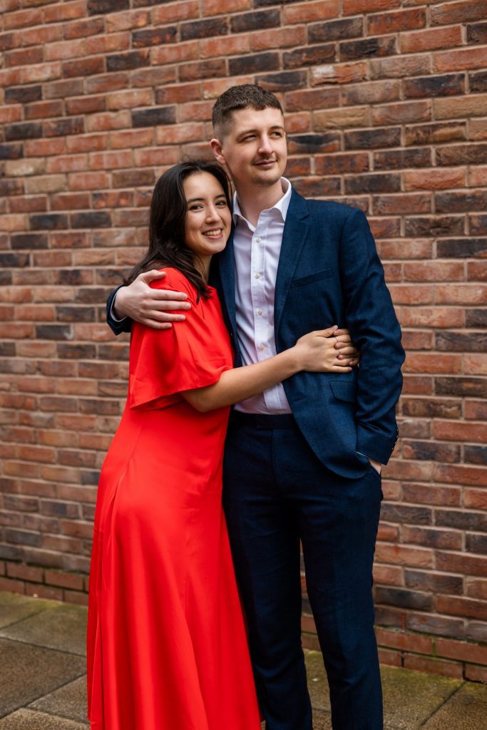 Engaged couple stood together leaning against a tall brick wall in Ancoats, Manchester - The bride to-be is wearing a vivid red dress, and the groom to-be is wearing a blue suit - Engagement Shoot - Isabelle B Photography