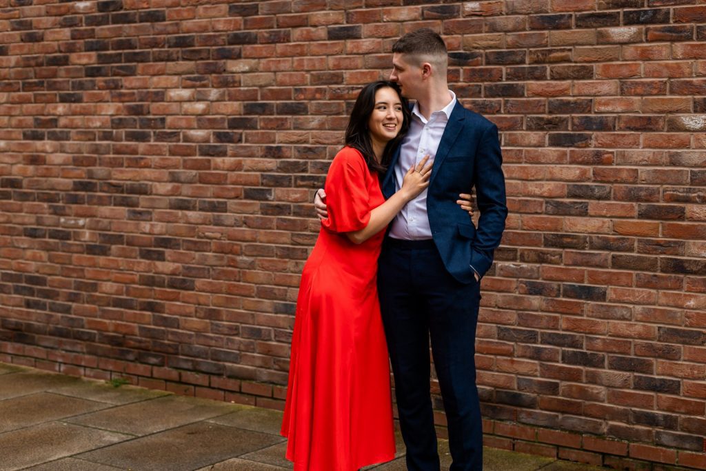 Engaged couple stood together leaning against a tall brick wall in Ancoats, Manchester - The bride to-be is wearing a vivid red dress, and the groom to-be is wearing a blue suit - Engagement Shoot - Isabelle B Photography