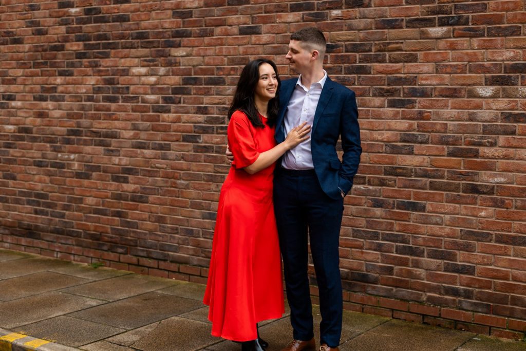 Engaged couple stood together leaning against a tall brick wall in Ancoats, Manchester - The bride to-be is wearing a vivid red dress, and the groom to-be is wearing a blue suit - Engagement Shoot - Isabelle B Photography