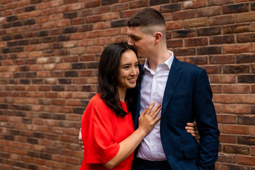 Engaged couple stood together leaning against a tall brick wall in Ancoats, Manchester - The bride to-be is wearing a vivid red dress, and the groom to-be is wearing a blue suit - Engagement Shoot - Isabelle B Photography