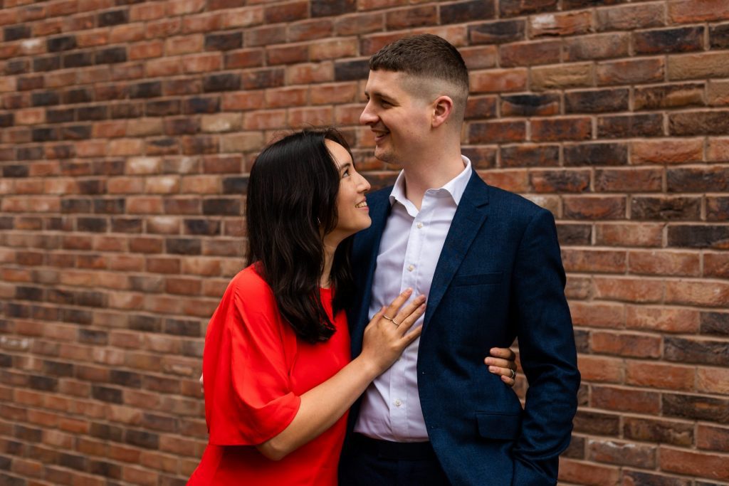 Engaged couple stood together leaning against a tall brick wall in Ancoats, Manchester - The bride to-be is wearing a vivid red dress, and the groom to-be is wearing a blue suit - Engagement Shoot - Isabelle B Photography