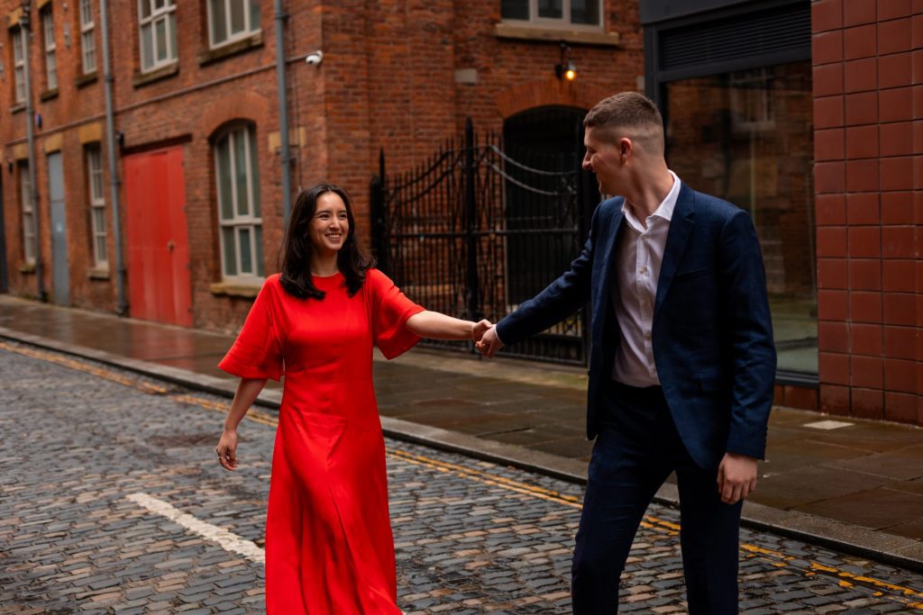 Newly engaged couple walking together in Ancoats, Manchester - The bride to-be is wearing a vivid red dress & the groom to-be is wearing a blue suit - Engagement Shoot - Isabelle B Photography