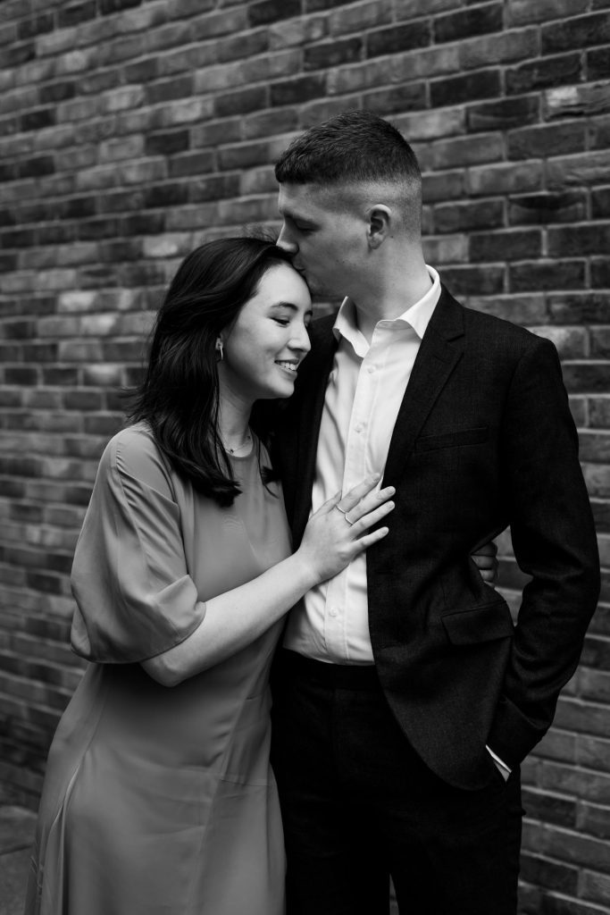 Engaged couple stood together leaning against a tall brick wall in Ancoats, Manchester - The bride to-be is wearing a vivid red dress, and the groom to-be is wearing a blue suit - Engagement Shoot - Black & White - Isabelle B Photography