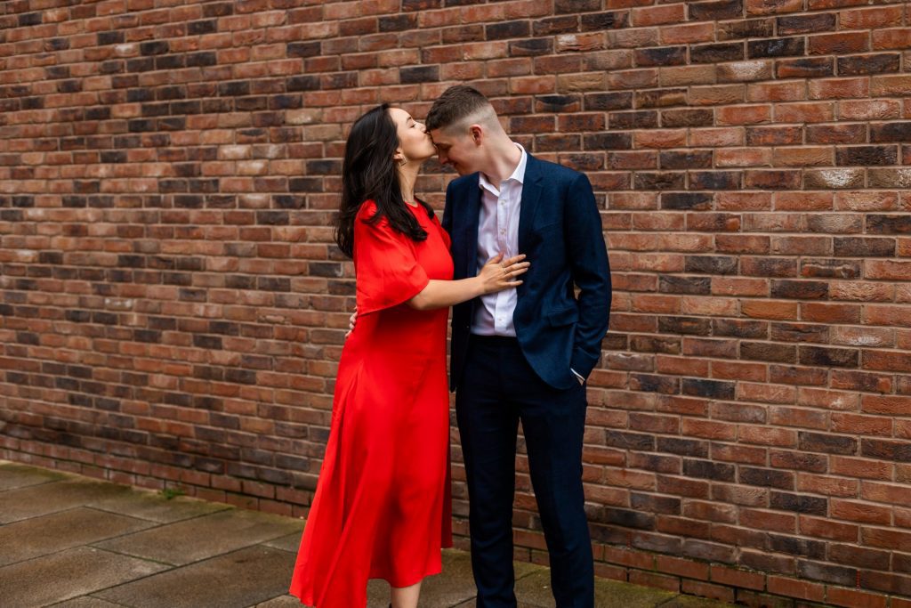 Engaged couple stood together leaning against a tall brick wall in Ancoats, Manchester - The bride to-be is wearing a vivid red dress, and the groom to-be is wearing a blue suit - The bride is giving her fiancé a kiss on the forehead - Engagement Shoot - Isabelle B Photography