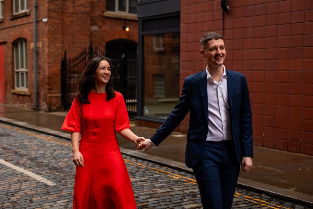 Newly engaged couple walking together in Ancoats, Manchester - The bride to-be is wearing a vivid red dress & the groom to-be is wearing a blue suit - Engagement Shoot - Isabelle B Photography