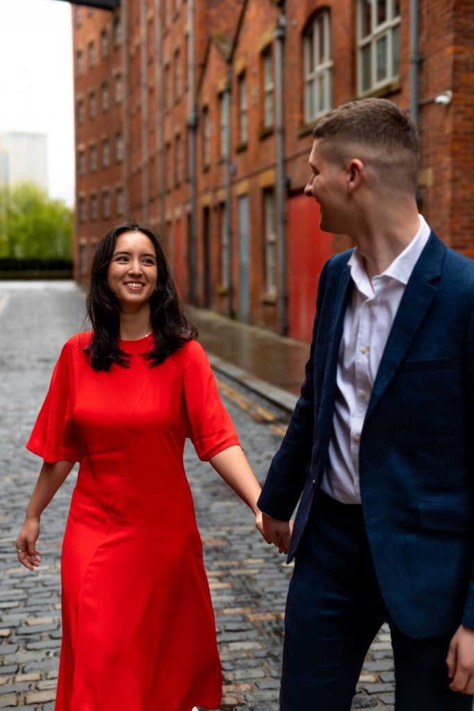 Newly engaged couple walking together in Ancoats, Manchester - The bride to-be is wearing a vivid red dress & the groom to-be is wearing a blue suit - Engagement Shoot - Isabelle B Photography