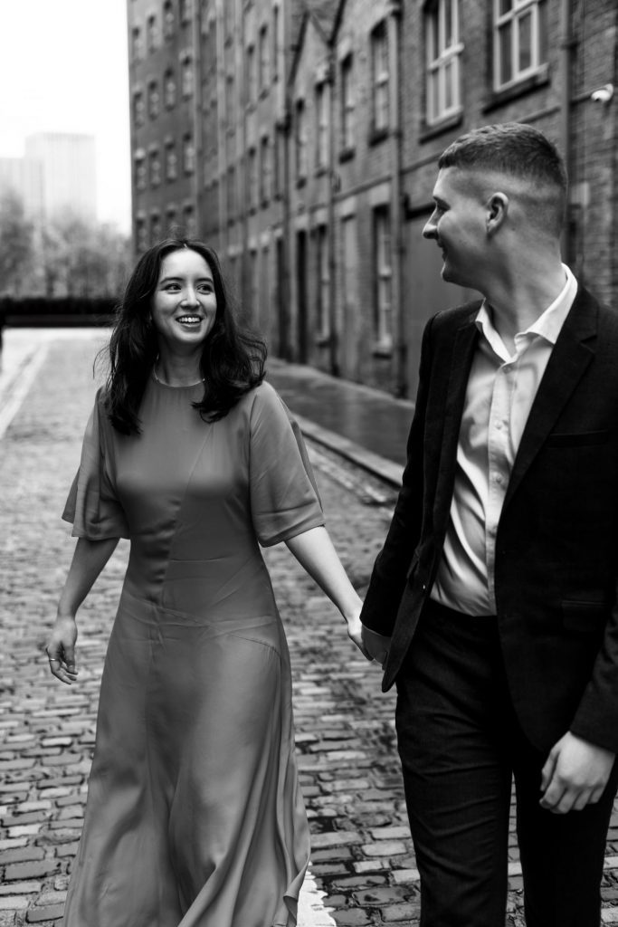 Newly engaged couple walking together in Ancoats, Manchester - The bride to-be is wearing a vivid red dress & the groom to-be is wearing a blue suit - Engagement Shoot - Black & White - Isabelle B Photography