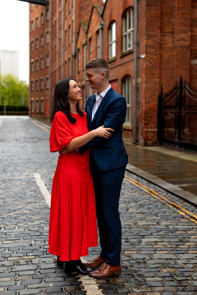 Newly engaged couple walking together in Ancoats, Manchester - The bride to-be is wearing a vivid red dress & the groom to-be is wearing a blue suit - Engagement Shoot - Isabelle B Photography