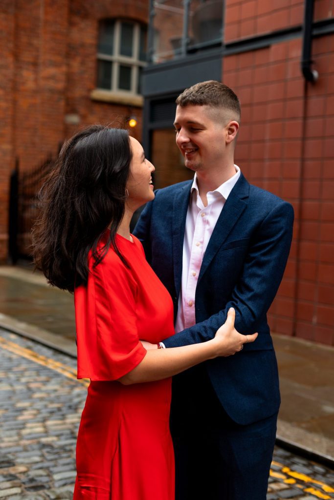 Newly engaged couple walking together in Ancoats, Manchester - The bride to-be is wearing a vivid red dress & the groom to-be is wearing a blue suit - Engagement Shoot - Isabelle B Photography