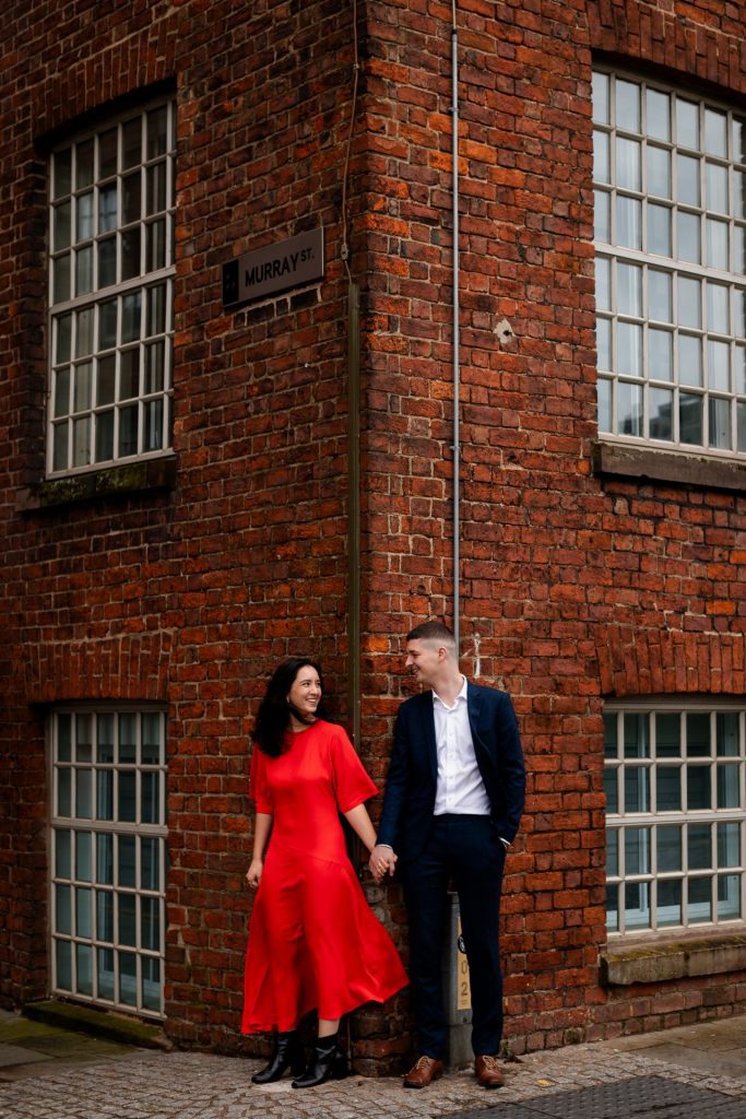 Newly engaged couple walking together in Ancoats, Manchester - The bride to-be is wearing a vivid red dress & the groom to-be is wearing a blue suit - Engagement Shoot - Isabelle B Photography