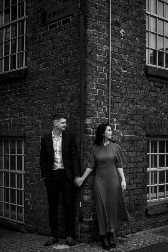 Newly engaged couple walking together in Ancoats, Manchester - The bride to-be is wearing a vivid red dress & the groom to-be is wearing a blue suit - Engagement Shoot - Black & White - Isabelle B Photography