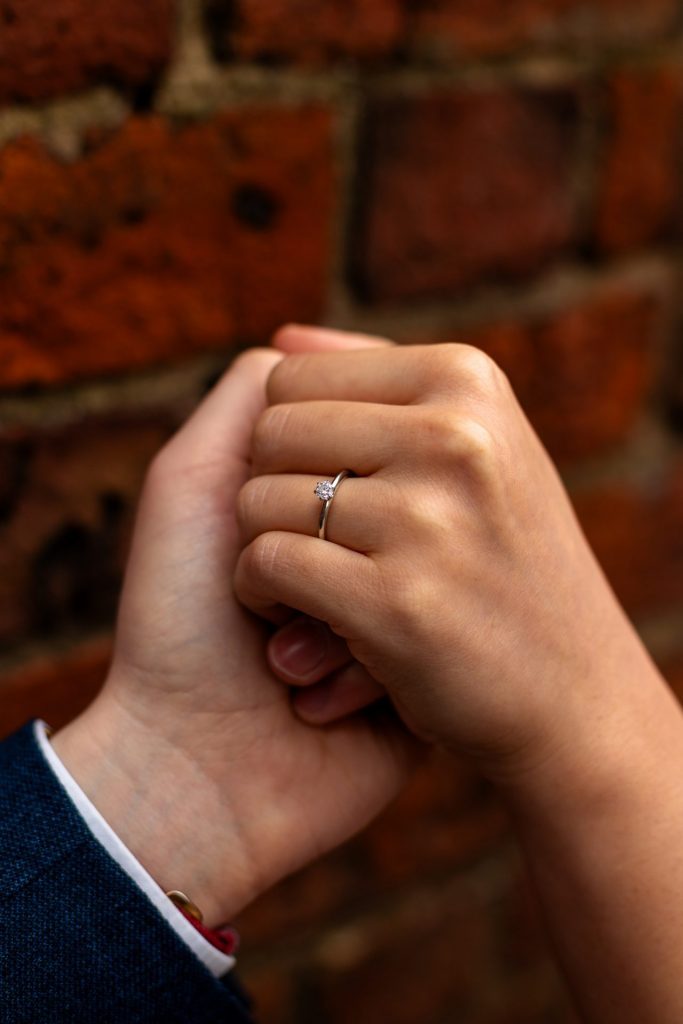 Close up of engagement ring on the bride to-be's hand again a red rick wall in Ancoats, Manchester - The bride to-be is wearing a vivid red dress & the groom to-be is wearing a blue suit - Engagement Shoot - Isabelle B Photography