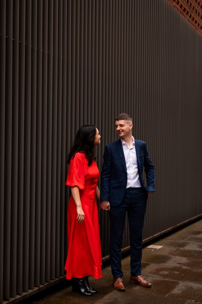 Newly engaged couple walking together in Ancoats, Manchester - The bride to-be is wearing a vivid red dress & the groom to-be is wearing a blue suit - Engagement Shoot - Isabelle B Photography