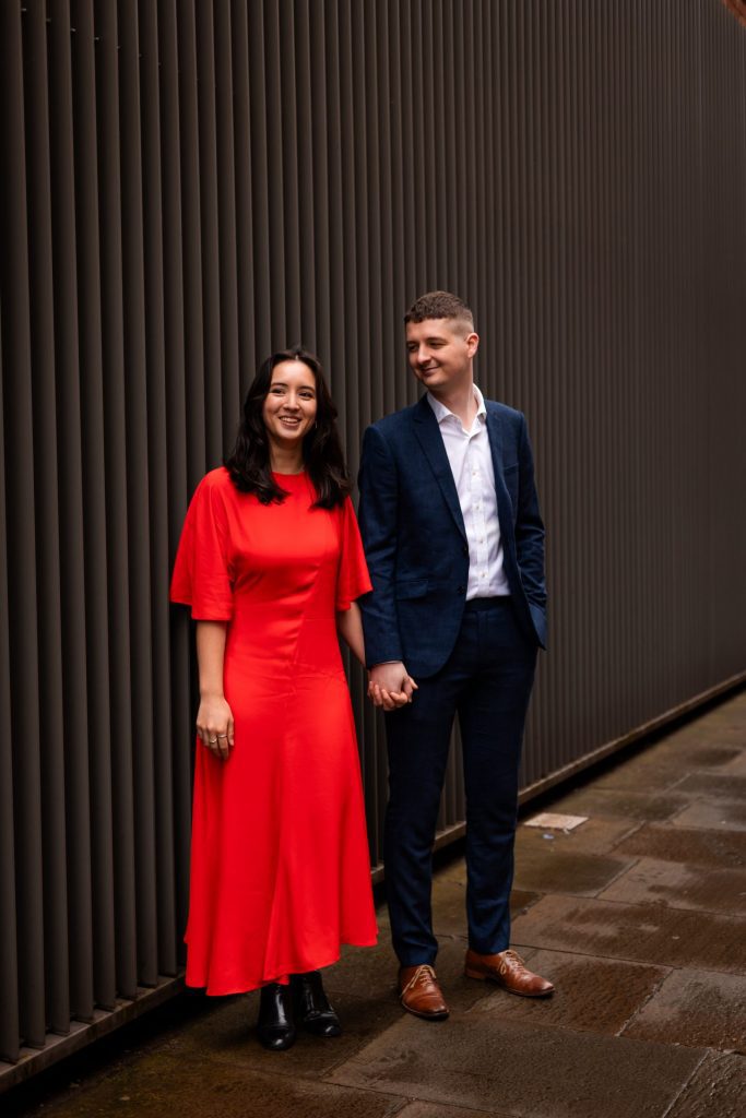 Newly engaged couple walking together in Ancoats, Manchester - The bride to-be is wearing a vivid red dress & the groom to-be is wearing a blue suit - Engagement Shoot - Isabelle B Photography