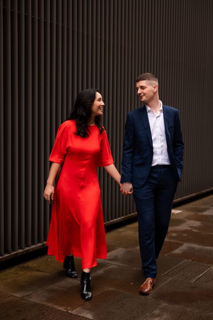 Newly engaged couple walking together in Ancoats, Manchester - The bride to-be is wearing a vivid red dress & the groom to-be is wearing a blue suit - Engagement Shoot - Isabelle B Photography