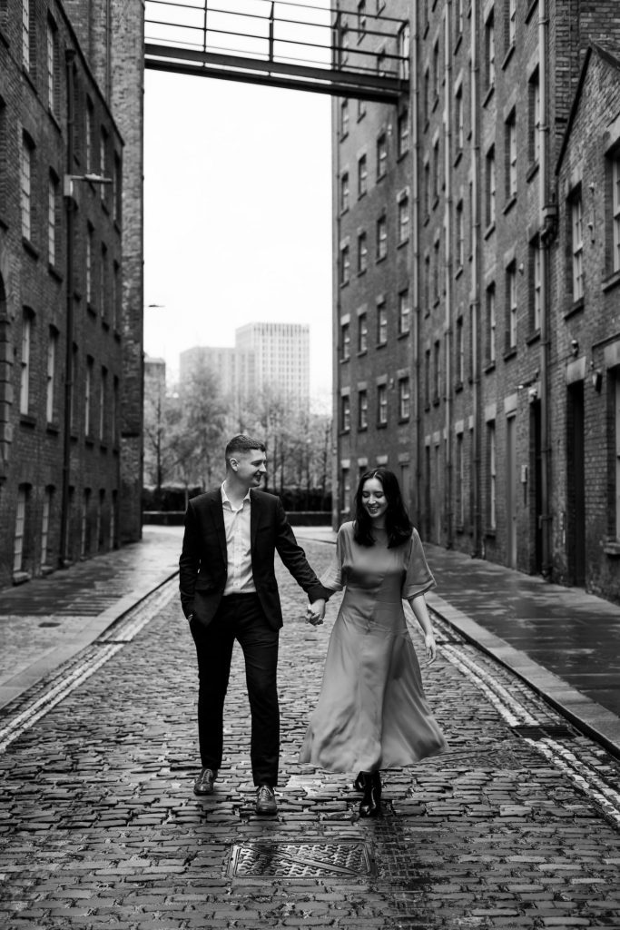 Newly engaged couple walking together in Ancoats, Manchester - The bride to-be is wearing a vivid red dress & the groom to-be is wearing a blue suit - Engagement Shoot - Black & White - Isabelle B Photography