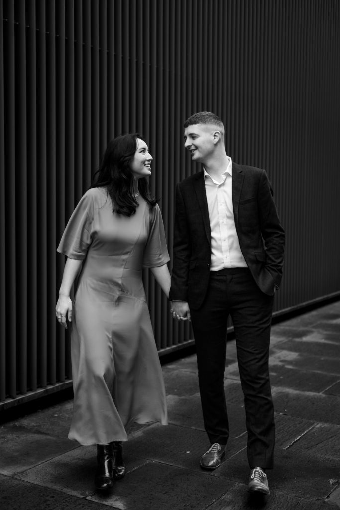 Newly engaged couple walking together in Ancoats, Manchester - The bride to-be is wearing a vivid red dress & the groom to-be is wearing a blue suit - Engagement Shoot - Black & White - Isabelle B Photography