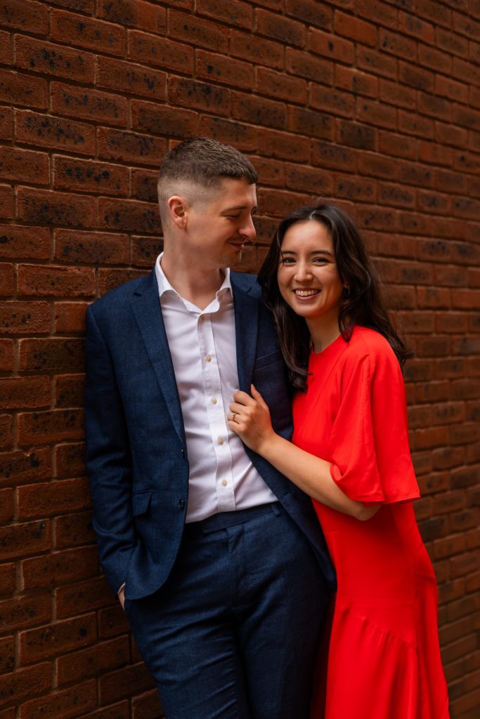 Newly engaged couple walking together in Ancoats, Manchester - The bride to-be is wearing a vivid red dress & the groom to-be is wearing a blue suit - Engagement Shoot - Isabelle B Photography
