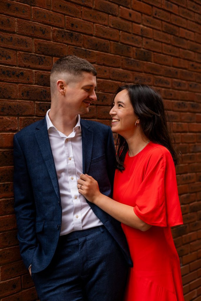 Newly engaged couple walking together in Ancoats, Manchester - The bride to-be is wearing a vivid red dress & the groom to-be is wearing a blue suit - Engagement Shoot - Isabelle B Photography