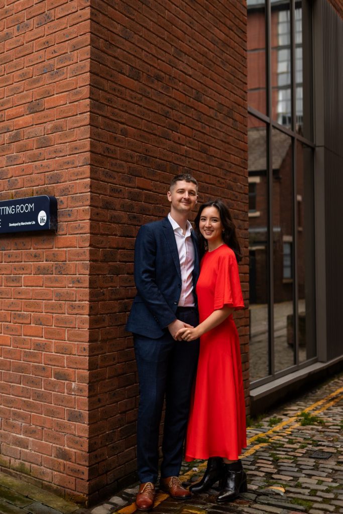 Newly engaged couple walking together in Ancoats, Manchester - The bride to-be is wearing a vivid red dress & the groom to-be is wearing a blue suit - Engagement Shoot - Isabelle B Photography