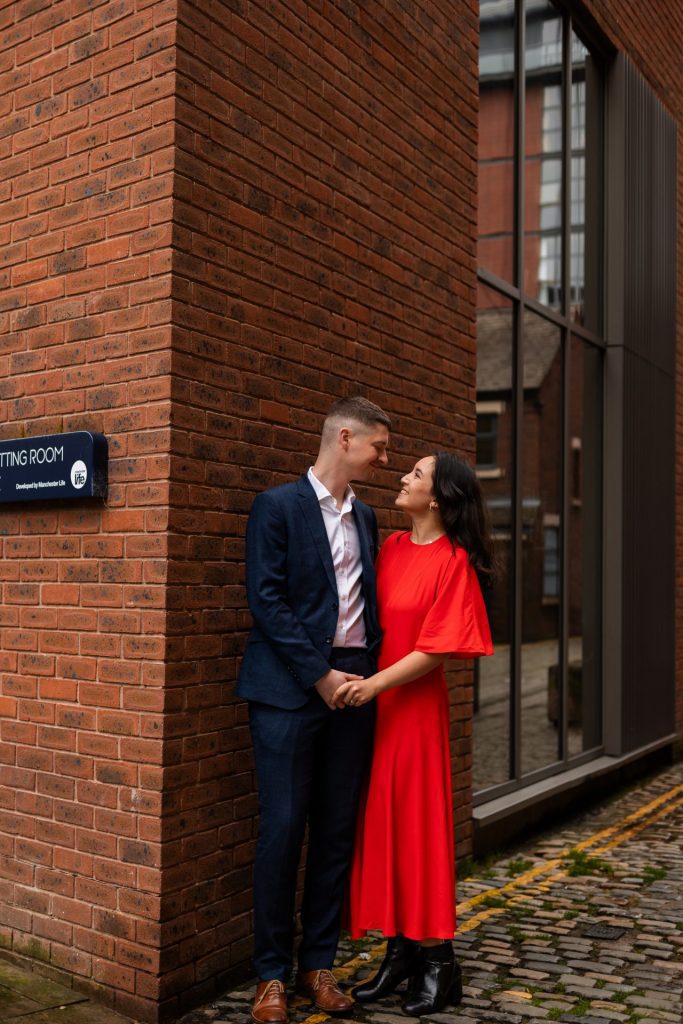 Newly engaged couple walking together in Ancoats, Manchester - The bride to-be is wearing a vivid red dress & the groom to-be is wearing a blue suit - Engagement Shoot - Isabelle B Photography